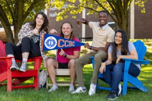 Four students sit in colorful chairs on a sunlit, grassy lawn with college buildings in the background. One holds a pennant reading "CCBC."
