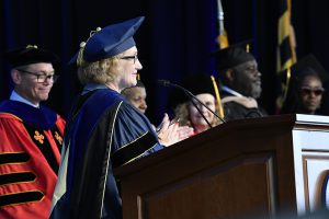 Woman in commencement regalia applauds at podium; others in commencement regalia appear in the background